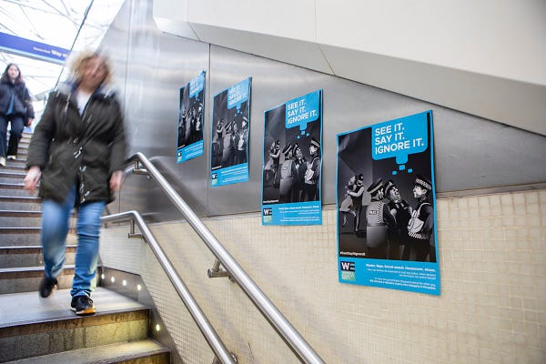 A member of the public walks down the stairs past four posters with black and white illustrations of police officers ignoring a woman being attacked by a fellow police officer. The words, "See it, say it, ignore it" are on the top right of the poster, and the poster is in a busy train station. 