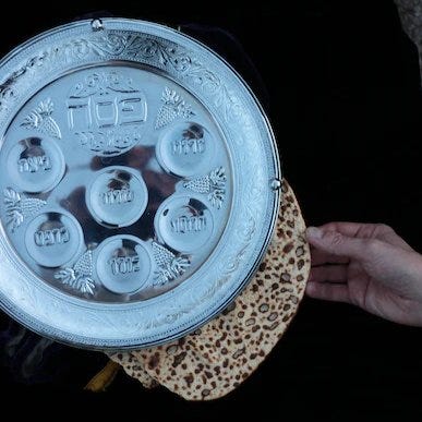 A Seder plate, Matzah and a hand against a black background.