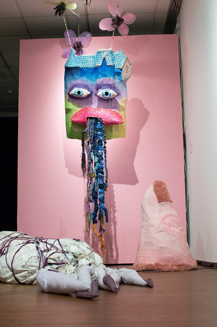 Installation shot of papiermache castle with eyes and mouth. From the mouth are multicolored yarn tendrils resembling roots, knots. A pink papiermache finger points at the castle. Fairies appear to hold the castle. The fairies have pastel wings and are in the shape of hands holding beaded string that connect to the back of the castle. 