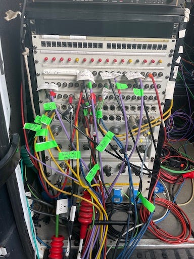1st photo: a patch bay on the back of a broadcast truck. Loads of wires going in and out, most of which have green or white bits of tape on them labelling their purpose. 2nd: Me plugging cables in at the back of another rack of kit.