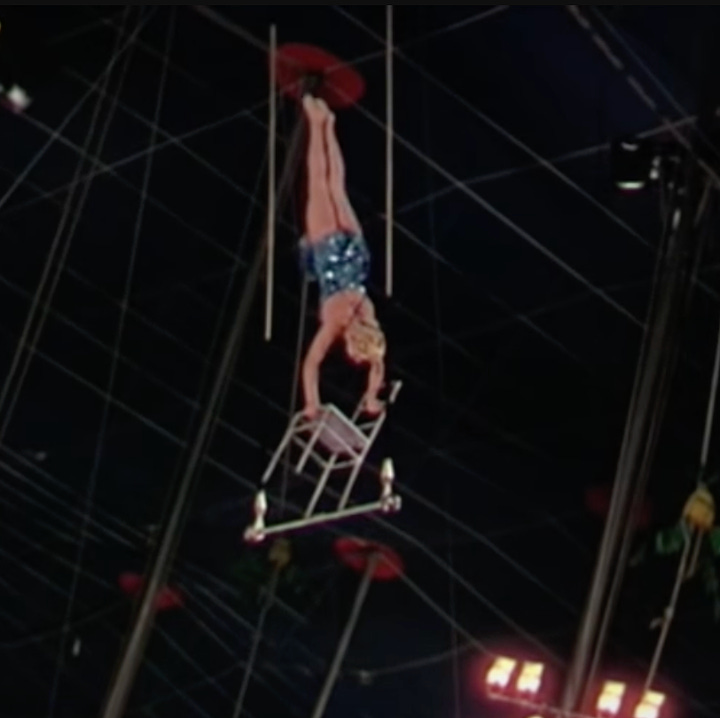 A man balances on a chair balanced on a swinging trapeze at the circus; actress Betty Hutton pulls off the stunning feat in the movie. 