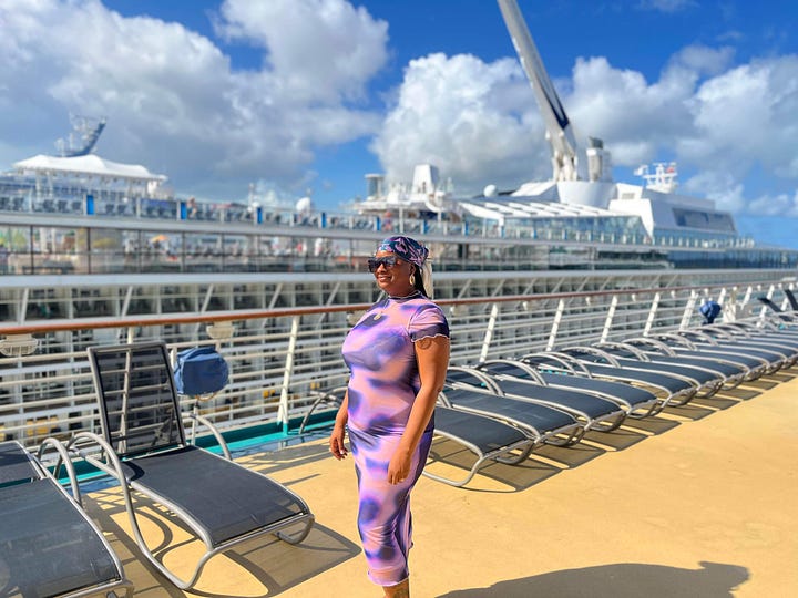 Black woman poses on a cruise ship in a multicolored purple dress. Black woman faces toward the Bahamian Ocean holding out the peace sign on each side.