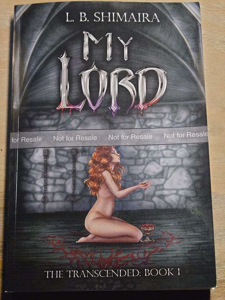 The background is that of a dungeon in grey and blueish hues.  In the upper part of the cover it reads L. B. Shimaira, and below that My Lord. The title lettering appears in a silver metallic font with red and purple blood dripping from the bottom. In the lower half of the cover a naked white woman with wavy red hair is seated on her knees, facing right. She has her hands clasped together in front of her and blood is spilling from them into a chalice by her knees. Her eyes are closed, and her chin is slightly turned up. The floor around her is covered in red blood that slowly changes to purple as it runs off-screen towards the viewer. To the left of her, on the wall, are shackles.  Below her is the text: the transcended, book 1.      Image description of the back cover:  The same background as that of the front cover is continued here (a dungeon in grey and blueish hues) however now there's a white man on his knees on the floor, facing the viewer. He's wearing dark leather leggings and is topless, his long black hair cascading over his muscular shoulders. His hands are outstretched to the side with his wrists chained, the chains going off-page left and right. He's smirking deviously and his chest is covered with cuts that bleed purple. The floor around him is covered with purple blood.  The text on the back reads:  "You try to comfort yourself with visions of events that didn't happen, but that doesn't mean that what did happen left no scars."  After losing hearth and home, Meya attempts to escape the traumatic memories by travelling east. In 13th century Tristanja, however, it’s not safe to be alone. A local slave trader catches wind of the kinless woman and abducts her in the dead of night. Beaten and abused, the now meek Meya is sold to Lord Deminas. He's known to be cruel, yet becomes strangely protective of his new chambermaid, punishing anyone who dares hurt her. After cutting her to drink her blood, he even uses his own to heal her wounds. Meya and her paramour wonder if Deminas' dark secret is why servants regularly vanish, including the lord's previous chambermaids. However, the two women quickly learn that Lord Deminas isn't the only danger lurking in the castle's shadows.  MY LORD is a queer, slow-burn erotic gothic horror novel about rediscovering yourself after trauma—with kinky, blood-drinking immortals and polyamory.