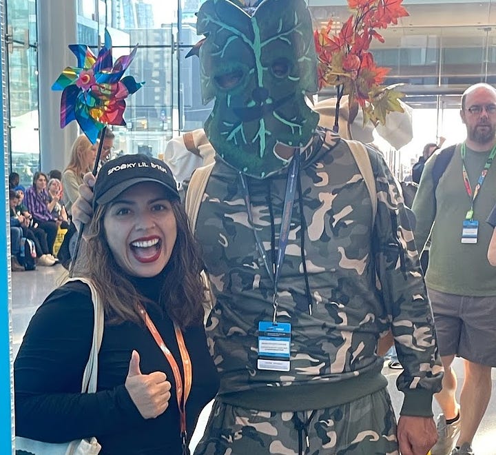 Lauren poses with a thumbs up next to a man cosplaying as a Korok in camo pants and Korok mask. Another man in Korok cosplay waving from an escalator.