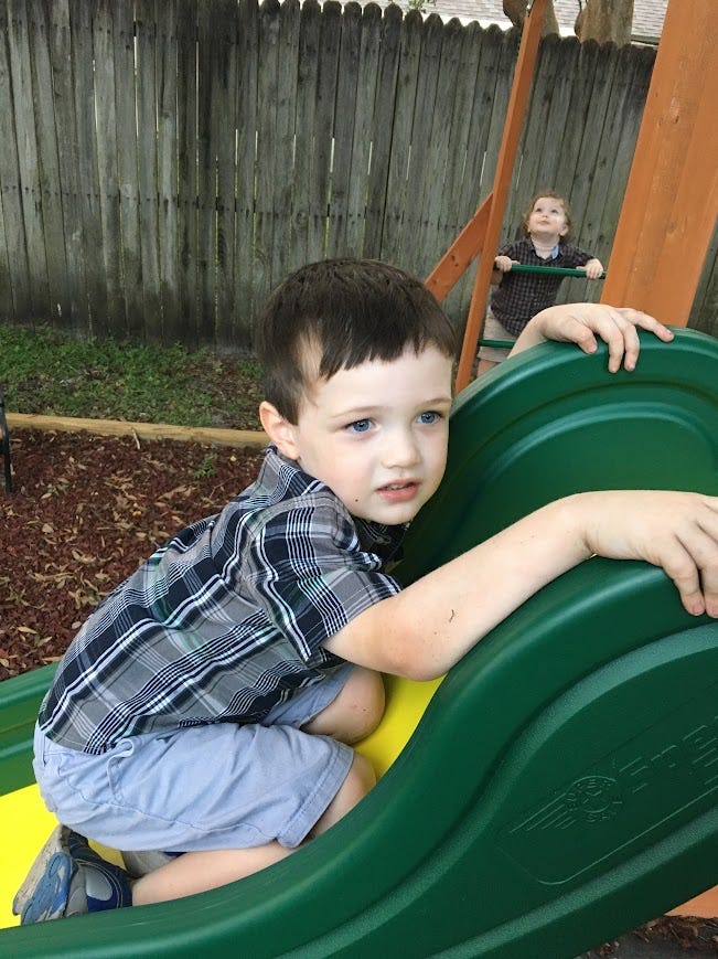 The image shows a young child standing on a wooden deck, gripping the rungs of a ladder that leads up to a playset. The child is looking upwards, perhaps at someone or something not visible in the photo. He is wearing a plaid shirt, shorts, and sandals, with a backdrop of a wooden privacy fence. The moment captured is tranquil, likely a snapshot of playtime or exploration. Second picture:  The image features a young boy sitting at the top of a green plastic slide, his hands gripping the sides as he prepares to slide down. He wears a plaid shirt and shorts, and his expression is one of concentration and cautious excitement. In the background, another child is visible, standing on a wooden deck. The setting suggests a playful and curious atmosphere in a residential backyard, with a wooden fence surrounding the area.