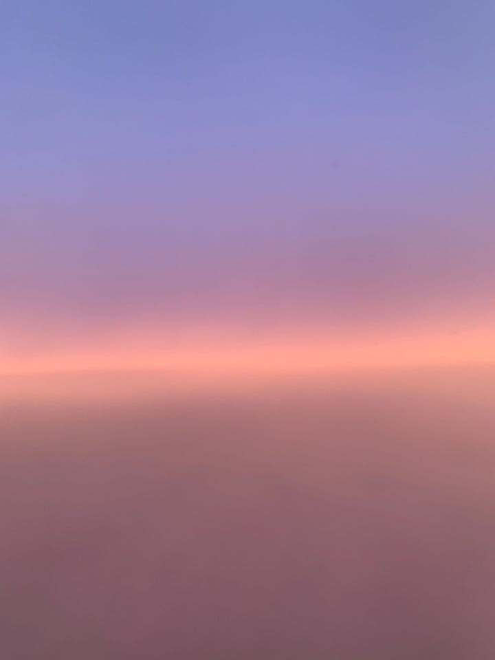 a blue sky with white clouds on the left; a picture from an airplane, of the place where the atmosphere and the horizon line meet, a mauve purple on the base of the image, a streak of peach pink in center, a line of deeper purple reddish, with blue purple on the top