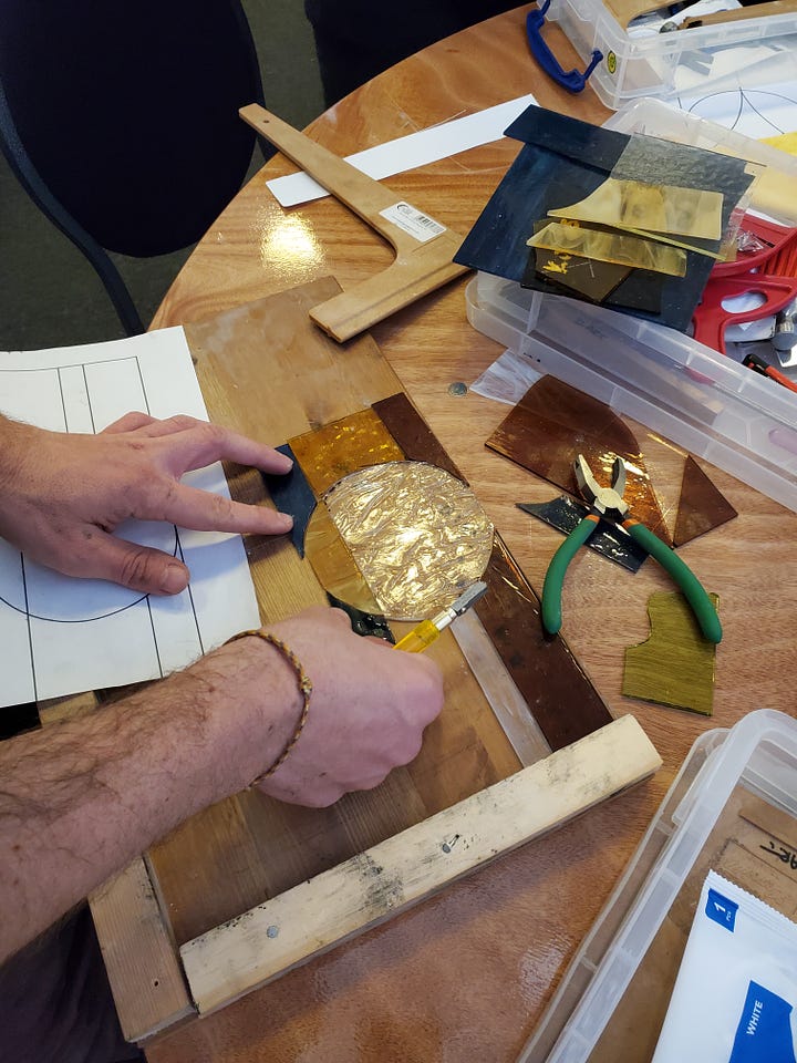 First Image: A person is working on a stained glass project. Their hands are carefully arranging pieces of colored glass, including yellow, gold, and black sections. Tools like pliers and a cutting tool are visible on the table, alongside various sheets of glass and a wooden frame. There is a paper design outline partially shown underneath the glass pieces. Second Image: A partially completed stained glass artwork is in progress, showing a colorful sunburst design. Green and yellow glass rays emanate from an orange half-sun, with blue and green glass pieces arranged at the bottom. The design is laid out in a wooden frame, with scattered glass pieces and tools like clamps, pliers, and a cutting tool visible around the workspace.