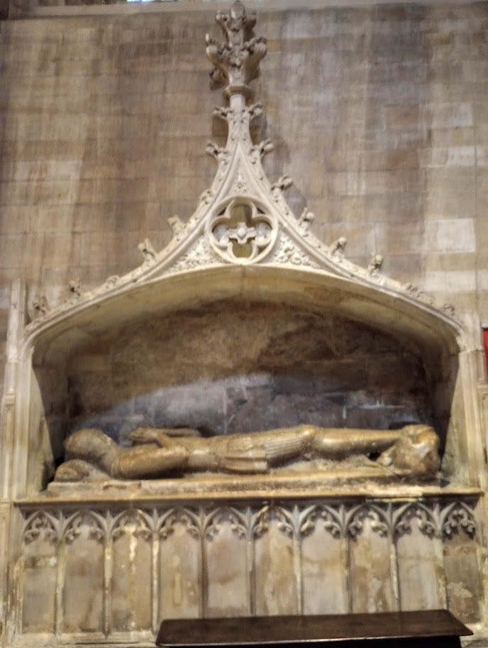 Tomb in wall, with canopy, featuring lifesize effigy of medieval knight with chainmail and helmet.