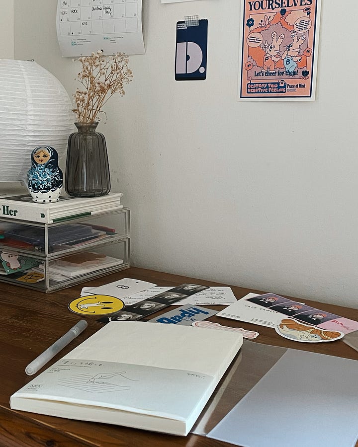 Gallery - Image 1: A messy desk featuring a new notebook with a blank cover, surrounded by stickers waiting to be added. / Image 2: A close-up of the open ruled notebook with stickers in the background.