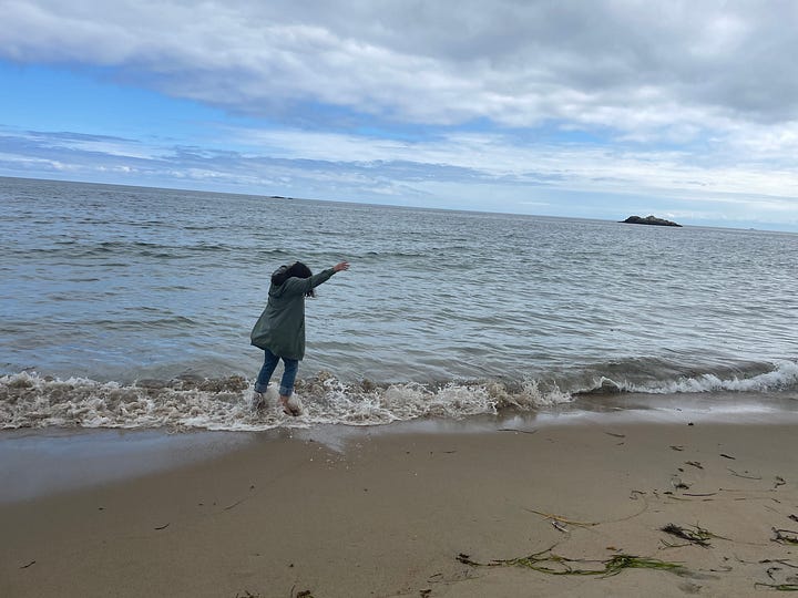 Two photos taken seconds apart. Jen is in jeans and a green sweatshirt-jacket. In one photo she is standing in the shallow white surf to her ankles only. In the next, she lifts her hands over her head as she tries to avoid the small shift in the water that had reached the bottom of her jeans.