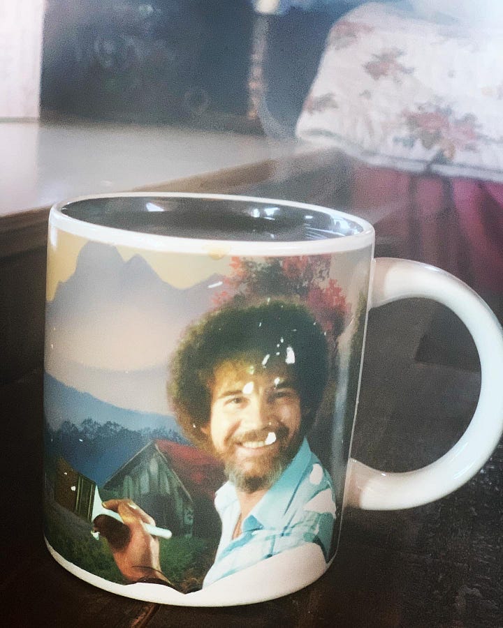 A mug, on one side a photo of Bob Ross with a paintbrush in hand, on the other, trees and and a landscape with the saying, "We don't make mistakes. We have happy accidents."