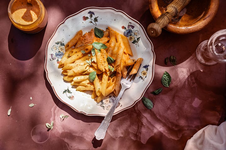 Creamy red pesto in a small glass jar on a red tablecloth, penne pasta with red pesto in an old fashioned pasta bowl with flowered edges on an outdoor table dappled with shadow from the trees and sunlight