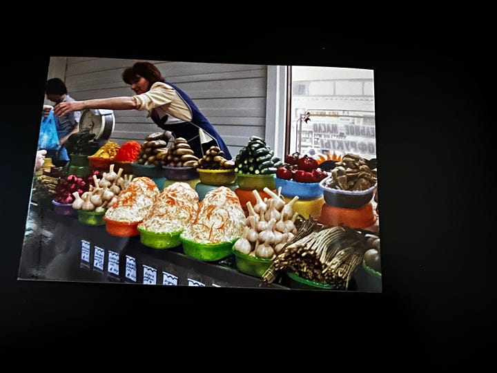 Two photos. Left to right: A woman in a blue apron and white shirt reaching over a countertop (two tiers) piled with bowls of pickled cucumbers and other kinds of pickles, pickled garlic, and sauerkraut. Second photo, a woman in a pink shirt and white apron, a kerchief tied over her hair, standing over a counter stall of radishes, dill, turnips, and other produce, mostly small bundles of leafy greens.