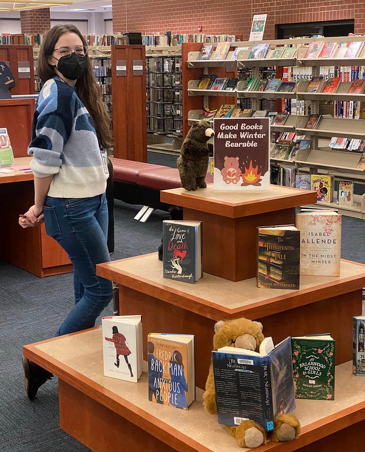 First image is a book display for women's history month featuring memoirs from female authors and the second is Marty posing next to a book display with the title "Good Books make winter bearable" with a plush bear posed nearby.