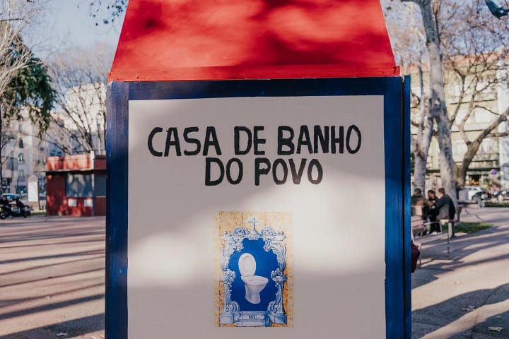 Foto 1: Un signore anziano, con indosso un cappotto scuro e un berretto, si trova accanto a un gabinetto pubblico bianco con un tetto rosso appuntito. Sul lato del gabinetto è presente una scritta in portoghese che recita "SANITÁRIO PÚBLICO INAUGURADO 2024 CONSTRUÍDO PELOS CIDADÃOS DE LISBOA", accompagnata da un disegno colorato che assomiglia a un sole sorridente. A sinistra del gabinetto ci sono due sedie di legno rosse e una bacinella verde. Lo sfondo mostra un ambiente urbano con alberi e edifici. Foto 2: Un cartello bianco con una sommità rossa e una cornice blu mostra la scritta "CASA DE BANHO DO POVO" in lettere nere. Sotto la scritta, c'è una piastrella bianca e blu che raffigura un gabinetto, con la scritta "WC" in piccolo in basso. Il cartello si trova all'aperto, con alberi spogli e persone sfocate sullo sfondo.