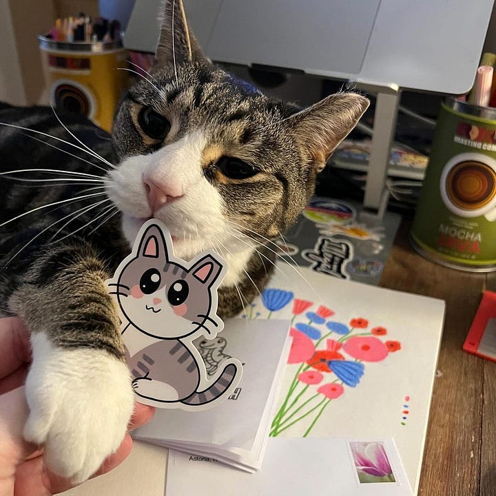 A grey and white tabby cat lounges on top of a sketch book and checks out the sticker of himself that his mom is holding. A black cat stands on top of a desk and checks out the sticker of himself that his mom is holding.