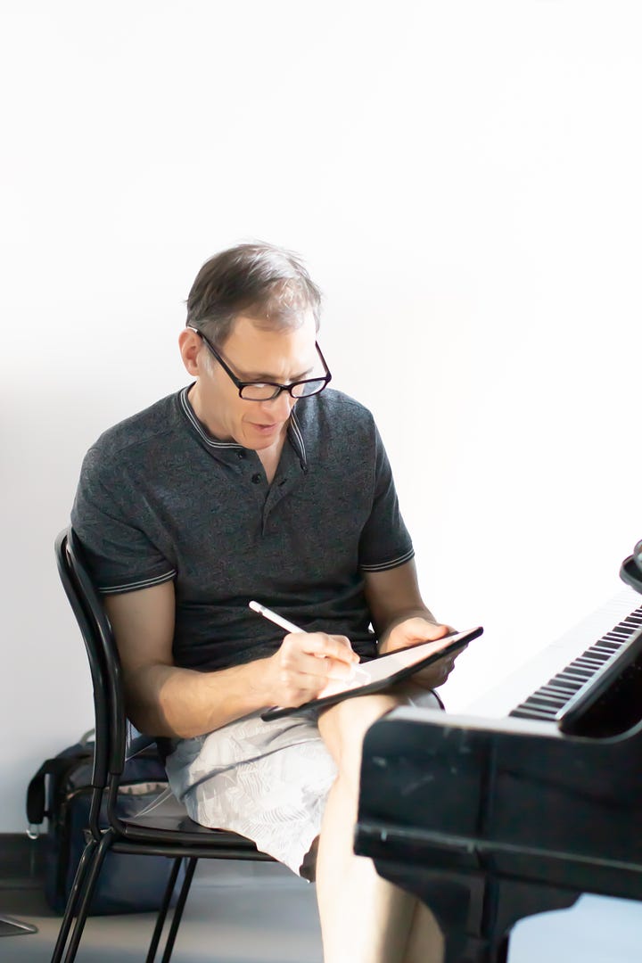 Susan Derry - woman in profile,  medium length brown bob and black sweater, hand on chin. editing the script in a rehearsal room. Howard Breitbart making notes on an iPad while sitting at a piano.