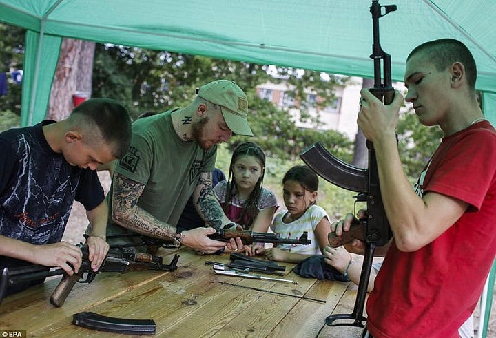 Pictures show young children being taught how to use rifles by Azov members with Swastika tattoos.