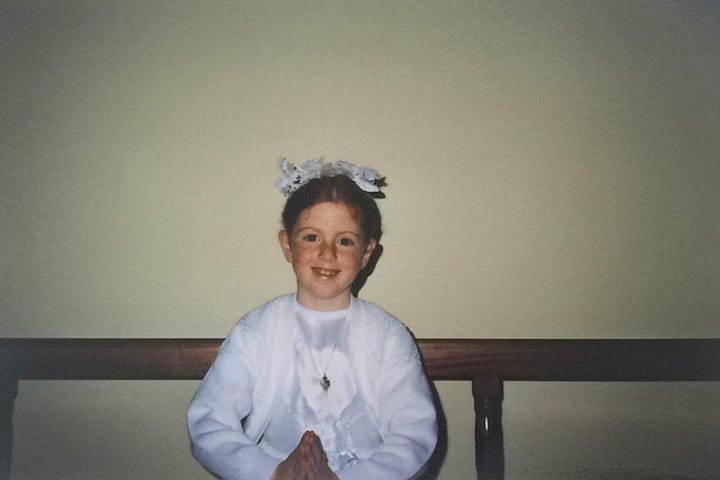 A young red-headed child making her first Holy Communion and standing next to a vase of droopy Daffodils in front of an image of Jesus Christ