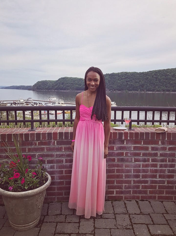 Left - Alexa is standing outside by the Hudson River in front of a half wall made of brick, at Shadows on the Hudson in Poughkeepsie, New York. There is a pot of pink flowers next to her. Her hair is in box braids that almost come down to her waist. She is wearing a floor length ombre pink gown with thin straps, and a hot pink wristband.   Right - Alexa is leaning against a brick wall outside of a restaurant in L.A. There is a thin plank of wood beside her, resembling a bar ledge. She is wearing a pink floor length gown that is almost the same color as the picture on the right. The straps of this dress are ruffled, and wider and thicker than the dress in the photo on the left, and the dress is made of a thicker material Alexa’s hair is still in box braids, but pulled back in a low ponytail.  Alexa is smiling at the camera in both photos. She is also wearing a necklace that comes down to the middle of her chest, in both photos. (The necklaces are different, but look somewhat similar.) 