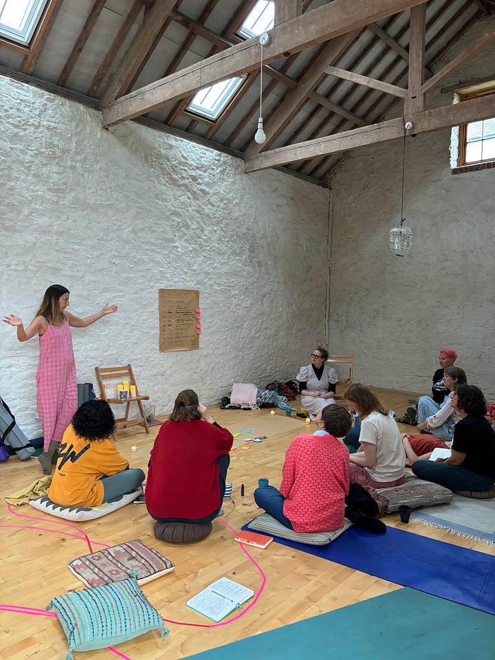 Sunset sky with a garden in the foreground, a blue shed to the right. A Black woman's hands holding a handmade cardboard loom with yellow wool. A floral mandala arrangement. A group of women sit in a circle in a barn. One woman is standing and gesturing widely (white, shoulder length brown hair, wearing a pink jumpsuit). A group of women sit in a circle lit only by firelight and candlelight