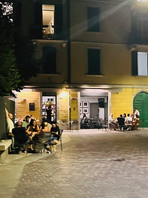 piazza full of tables and people, cobblestone streets, Via Vitani, yellow Italian stucco buildings with green doors, white Christmas lights in summer.