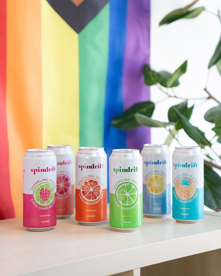 Two images side-by-side, each of six cans of spindrift. In the first, the colors of the cans are, in order, pink, peach, orange, green, blue, turquoise. In the second, the colors are, in order, pink, orange, peach, green, turquoise, purple. The purple can is inexplicably smaller. Behind each there is a progress flag, and the second picture has a letter board that said "happy pride" in white on a light pink background.