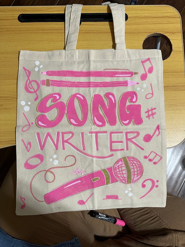 Image 1: A canvas tote which reads "Reader reader reader" in various hand lettered pink fonts and with illustrated books on the bottom; image 2: "Songwriter" hand lettered in pink acrylic paint and surrounded by music notes and a pink microphone; image 3: "Singer" lettered thee times in pink and surrounded by music notes and stars; image 4: "filmmaker" hand lettered in orange with a film strip over the top and a ribbon under the words that read "A Film By Micah Ariel Watson"