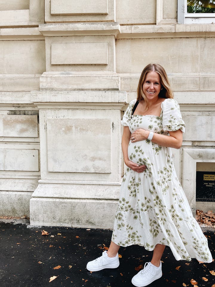 a woman stands in front of an off-white building. she is wearing a cream colored dress with green flowers and white sneakers, and is touching her baby bump and smiling at the camera. 