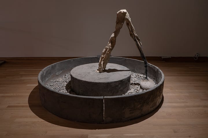 Two images of the sculpture. The image on the left shows the whole sculpture which includes a bony arm shaped like an upside down V. The arm is attached to a heavy stone by a twisted braid of hair. the stone is being dragged in a circle in a cement trough. On the right is a close up of the bony arm.
