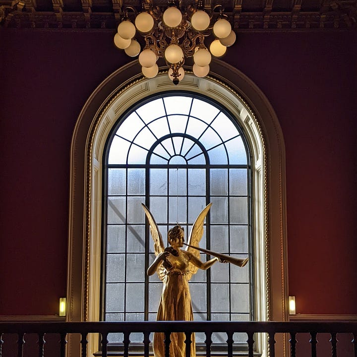 A group of four images in a museum; the top left of a golden angel statue before a window in a stairway; the top right of a swirling blue-and-yellow glass installation suspended from an ornate Corinthian ceiling before a tall archway; the bottom left of a statue in which a woman carrying a shield and staff is poised on top of a man's back, who is crushed underneath a second shield; the bottom right of another statue, this time of a woman sitting atop a marble wall, whose hand is being eaten by a man positioned underneath her on the ground.