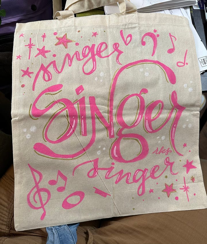 Image 1: A canvas tote which reads "Reader reader reader" in various hand lettered pink fonts and with illustrated books on the bottom; image 2: "Songwriter" hand lettered in pink acrylic paint and surrounded by music notes and a pink microphone; image 3: "Singer" lettered thee times in pink and surrounded by music notes and stars; image 4: "filmmaker" hand lettered in orange with a film strip over the top and a ribbon under the words that read "A Film By Micah Ariel Watson"