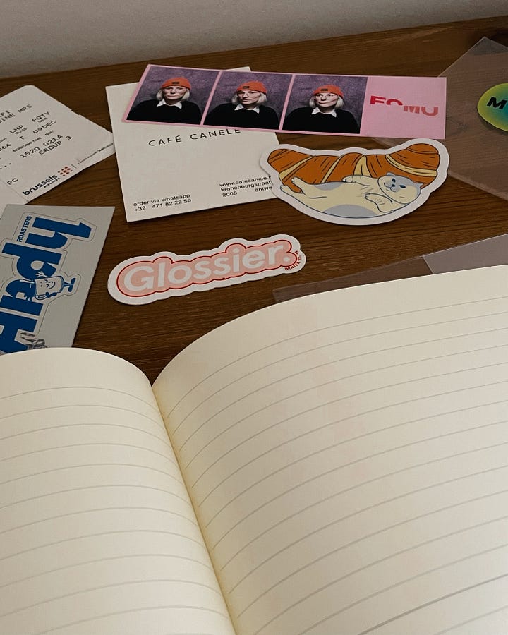 Gallery - Image 1: A messy desk featuring a new notebook with a blank cover, surrounded by stickers waiting to be added. / Image 2: A close-up of the open ruled notebook with stickers in the background.