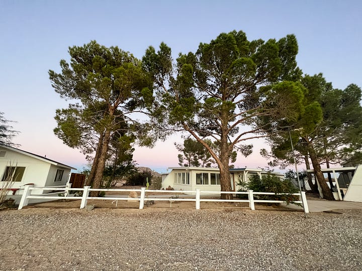 Selfie image of woman smiling and holding up a digital camera; image of a laptop and water bottle and coffee atop a low wooden coffee table with seating pillow; image of early morning sunlight streaming into a bedroom and adjacent bathroom; front facing view of three white buildings with large pinyon pine trees in foreground and a sunset sky of blues, pinks and purples in the background.