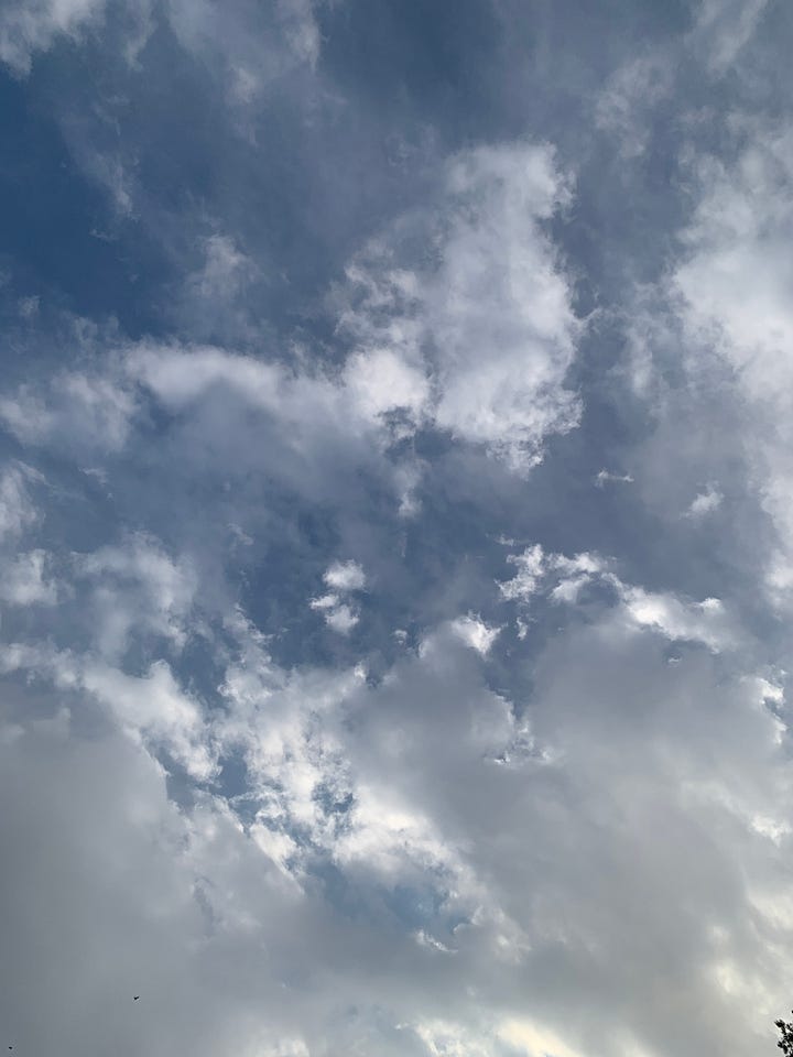 a blue sky with white clouds on the left; a picture from an airplane, of the place where the atmosphere and the horizon line meet, a mauve purple on the base of the image, a streak of peach pink in center, a line of deeper purple reddish, with blue purple on the top