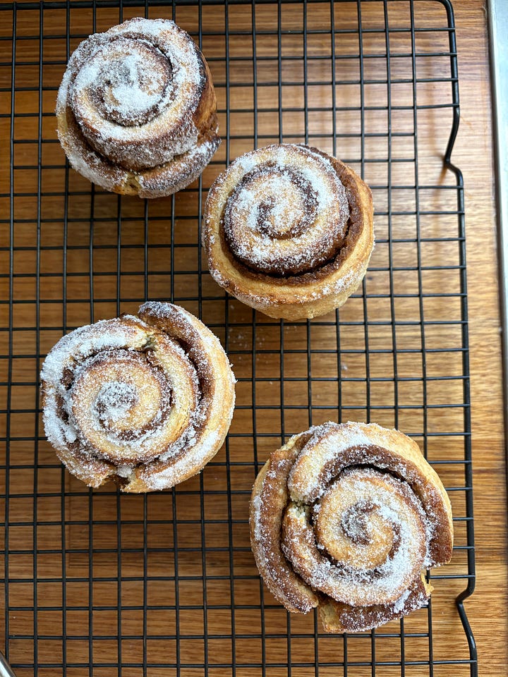 Four sugar-topped cinnamon scrolls on a wire rack. A cinnamon scroll on a plate with sliced kiwi fruit and an iced latte