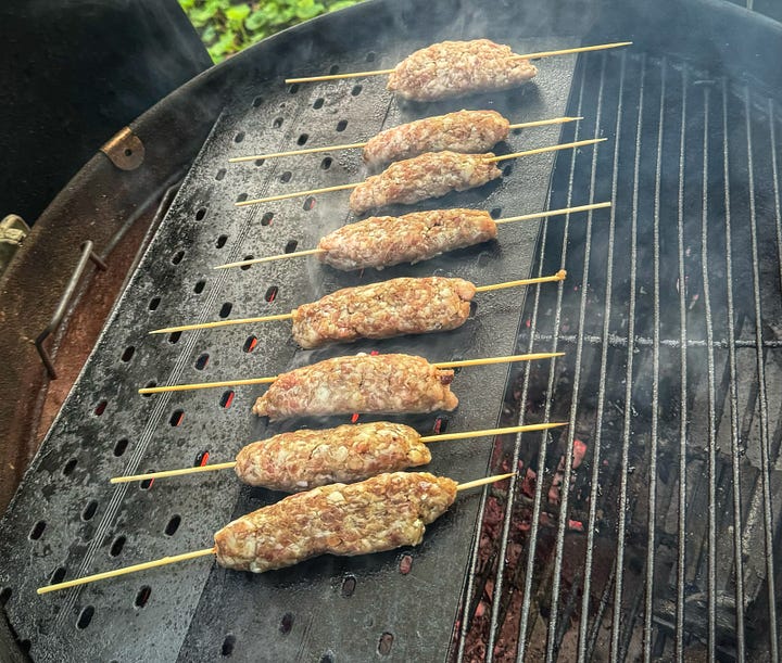 One photo of the skewered sausages cooking on a metal flat-top griddle over a charcoal grill. And a photo of the finished, browned skewered sausages piled on a plate, topped with a single, very small mint leaf. 