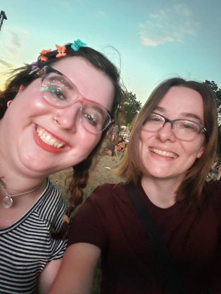 Holly, non binary person smiles while wearing a striped shirt glasses, butterfly clips and glasses with Janet, also smiling, wearing a black shirt and glasses. Janet stands wearing a blue and yellow patterned shirt and black pants stands next to Holly wearing a white patterned dress, a green wig, and antler flwer crown.