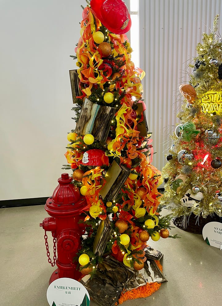 Christmas trees decorated with book characters and elements. On the left, a Christmas tree decorated with characters from Dr. Seuss's Cat in the Hat. On the right, a Christmas tree with a red fire hydrant nestled against it on the left. Decorations on the tree, in reds and yellows, include a red fireman's hat and yellow and orange ribbons intended to look like flames. The book is Fahrenheit 451 by Ray Bradbury. 