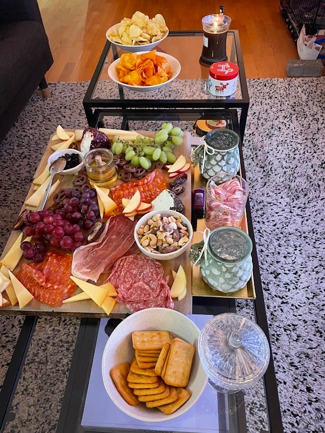 charcuterie board with many meets, crackers, cheeses,etc; embroidery project on table that shows a 'M' with a wreath around it