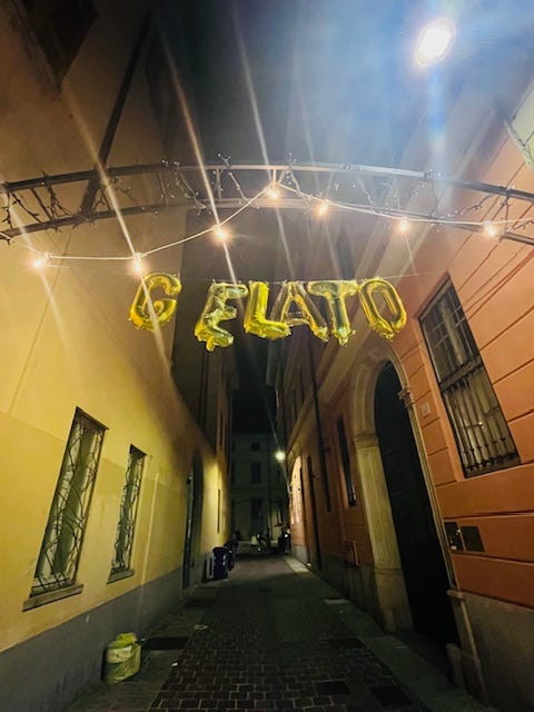 piazza full of tables and people, cobblestone streets, Via Vitani, yellow Italian stucco buildings with green doors, white Christmas lights in summer.