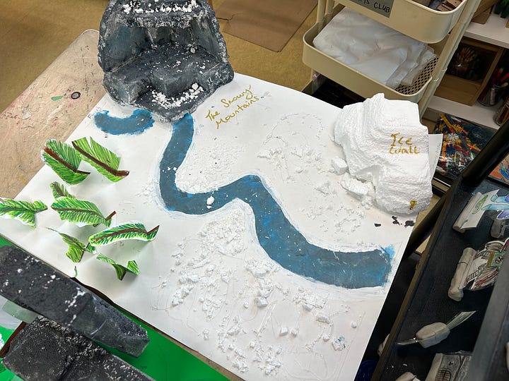 Square cardboard maps are decorated with paint, carved styrofoam, cardboard towers and paper cutouts to depict fantasy environments.