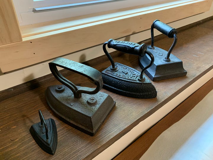 Left, Two long narrow paperweights, one a stainless steel bar with a knob handle and one in the shape of a branch with a smaller branch coming off it as a handle; right, four antique irons made of cast iron, one in miniature