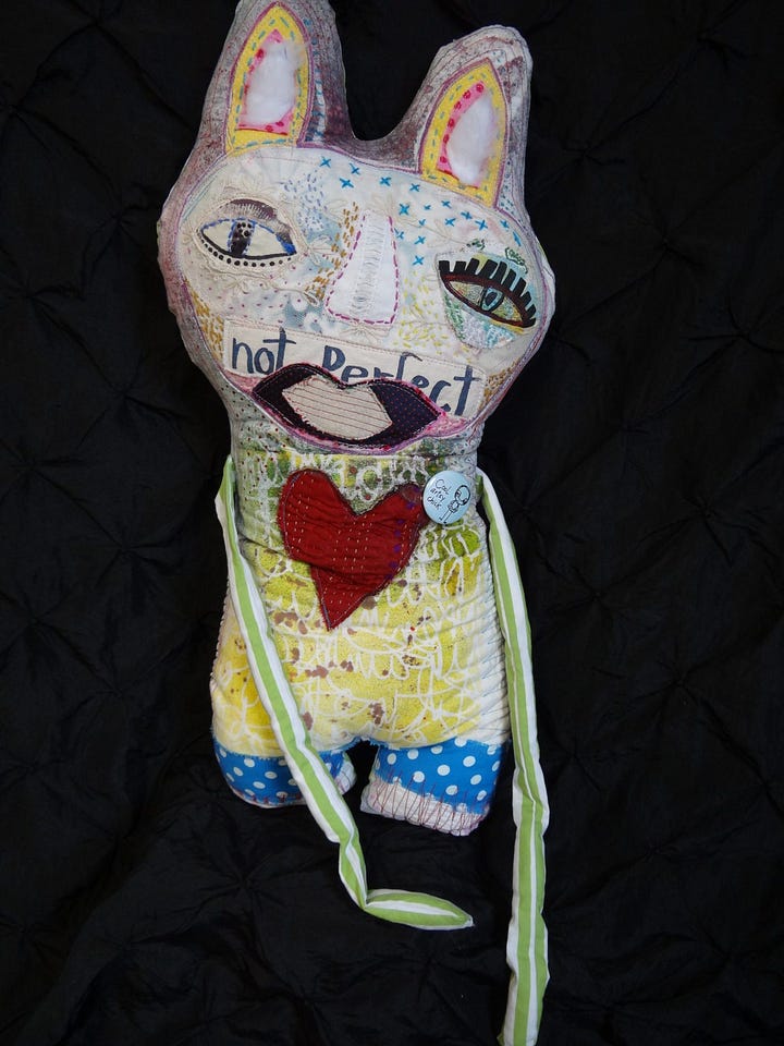 'Clockwise from top left, a plush doll with a brown face, a purple body and multicolored (primarily purple, pink and orange) feathers; the "not perfect" doll features a rabbit-eared figure with "not perfect" written over their mouth, they're sporting a yellow jumper with a big crimson heart on it; bottom, right, is a visual painting, "life," featuring two figures in Deveney's evocative "drip-like" style; and last, a doll with a bowling-pin-like body, with big sympathetic dark eyes, two big low ears that begin where earlobes would and a multicolored jumper on the body with knitted elements and a big red heart.
