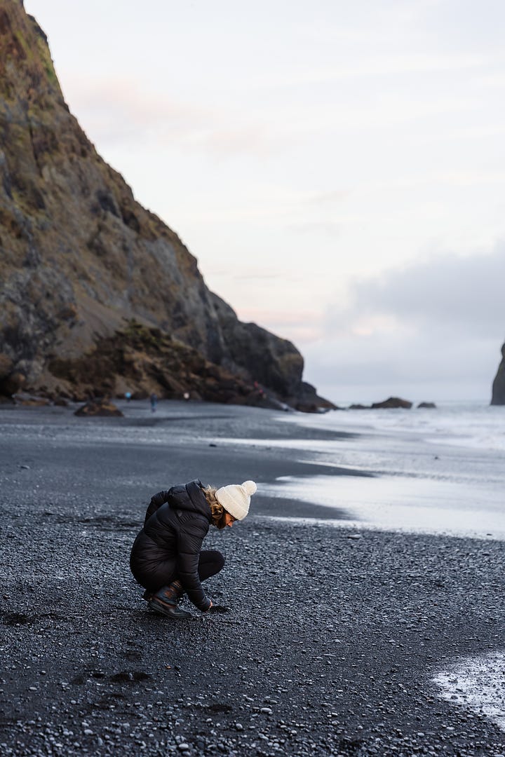 Photo of woman standing along a black sand beach to look at shells and pebbles; woman smiling sitting on a large boulder along a river filled with glacial melt and icebergs with a bridge in the distance; woman laughing and talking at an outdoor picnic table with a group of friends having coffee; selfie photo of two woman smiling wearing sunglasses and winter clothes.