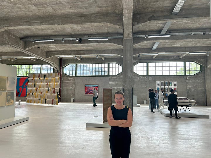 In the photo on the left, two women stand in front of a painting against a concrete wall. In the photo on the right, Katherine, wearing black, crosses her arms while standing in the industrial-style museum