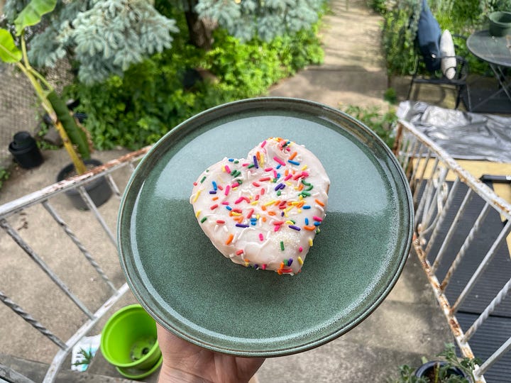 First photo of Lyss against a backdrop of red light, wearing black lipstick, a black choker, and a black tank top. Second photo of Kraftwerk live in concert, against a red background. Third photo of a donut with rainbow sprinkles. Fourth photo of a pitbull with a donut toy in her mouth, laying happily on a bed.