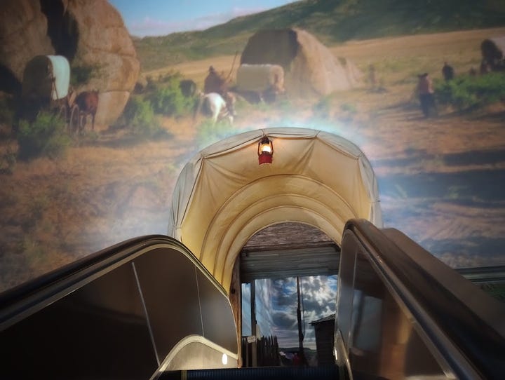 Long elevator with tableau of dry sand and rocks on either side, and a desert view of wagons at top. Indian woman in traditional dress with non-Native man in beard, buckskin, and carrying a rifle, man in wide-brimmed hat clambering up rocky hillside, followed by woman in dress and bonnet. Close view from top of escalator reveals that the passage at top imitates walking through a covered wagonn