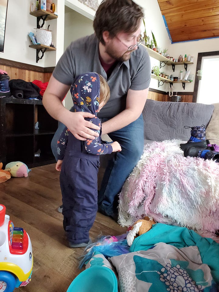 In the first image, my husband had his hands on Oren's shoulders as he just finished getting his snowpants on. In the second photo, Sybil is making a silly face at the camera while my husband puts Oren's shoes on in the background.