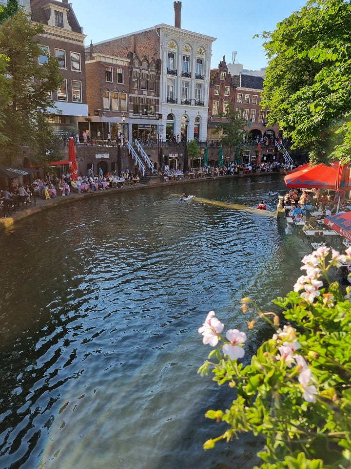 Photo 1: Canal in Utrecht showing people sitting outside enjoying bars and restaurants directly next to the canal. Photo 2: Dave looking up a the ceiling of the library which is domed and has circular light fixtures.